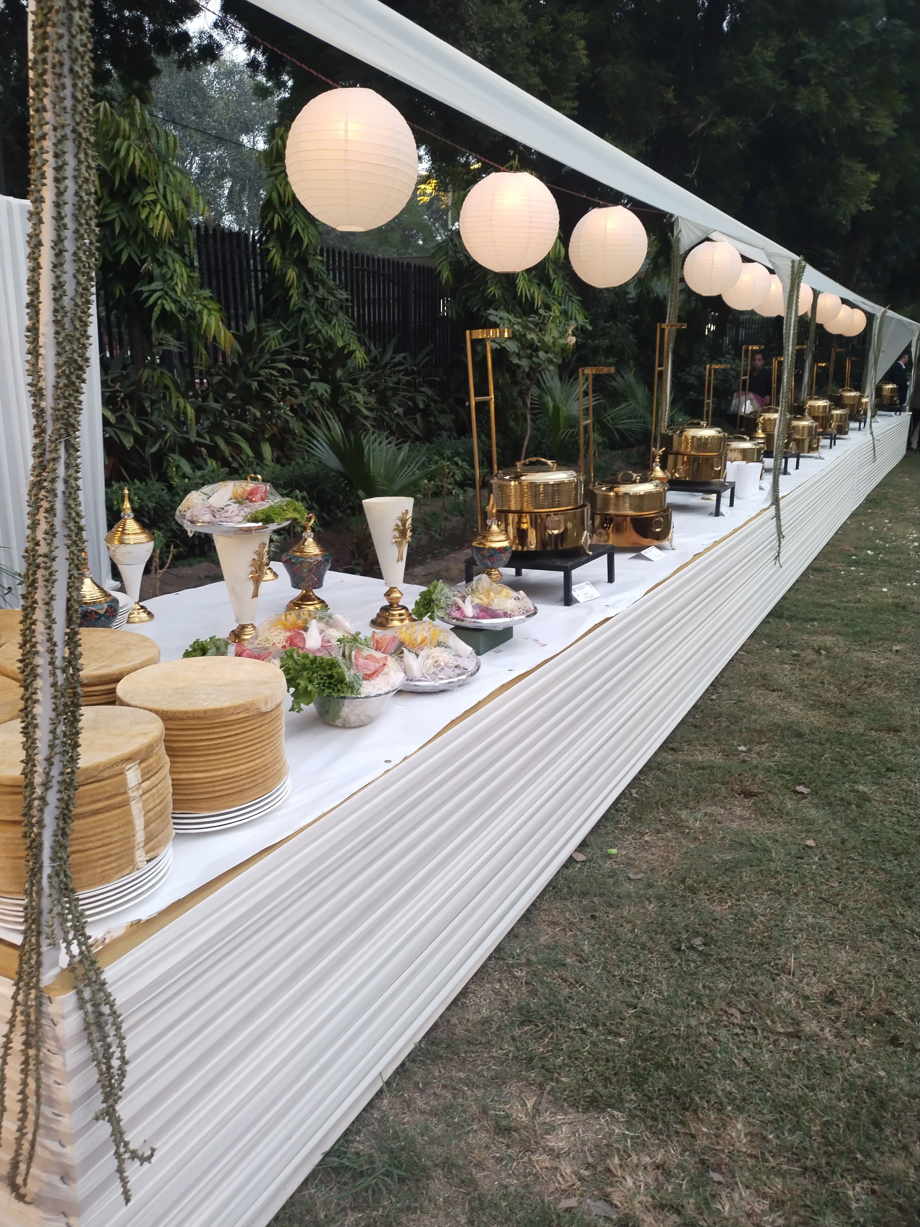 Fluted white live gourmet buffet counter with brass and gold chafing dishes and paper lanterns